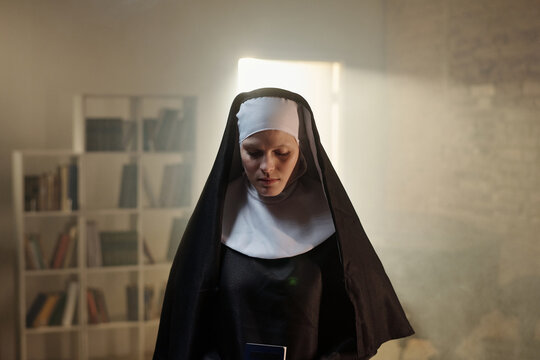 Caucasian woman dressed as nun standing in prison library holding book, looking down with serious expression, bookshelves filled with books visible in background