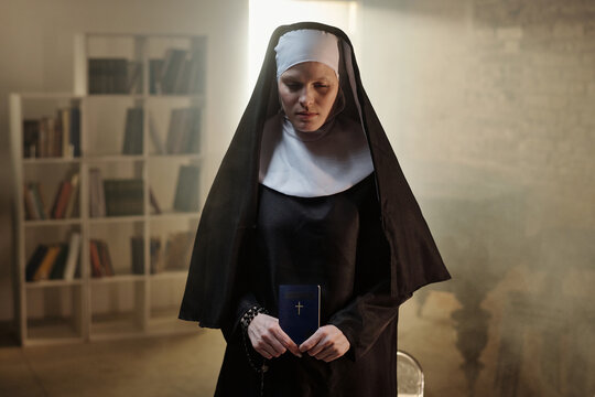 Caucasian woman dressed as nun standing with eyes closed holding prayer book and rosary in hands in prison library setting with bookshelves in background