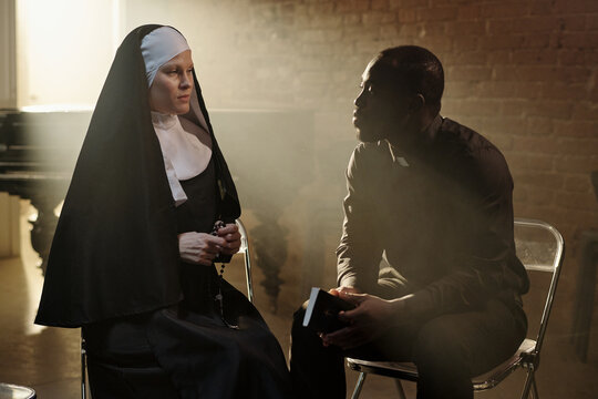 Caucasian nun sitting beside Black priest holding book, both engaged in conversation in prison library setting, sunlight streaming through window in background
