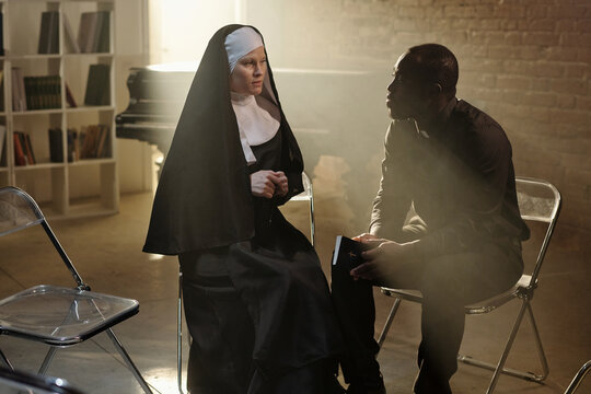 Caucasian nun sitting and talking with Black priest holding book in prison library setting, both engaged in serious conversation with shelves of books in background