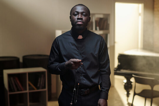 Portrait of Black priest standing in prison library holding rosary beads, looking directly at camera with serious expression, bookshelves and piano in background