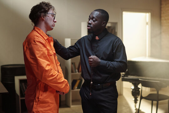 Caucasian young adult man wearing prison uniform standing in library while Black priest comforting him, both engaging in serious conversation near bookshelves in prison setting