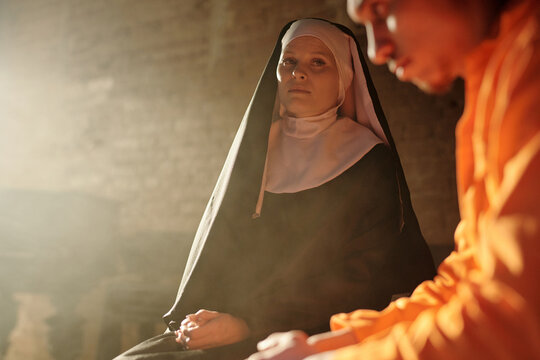 Caucasian  nun sitting beside young adult man wearing prison uniform, both inside prison library, nun looking forward while man looking down