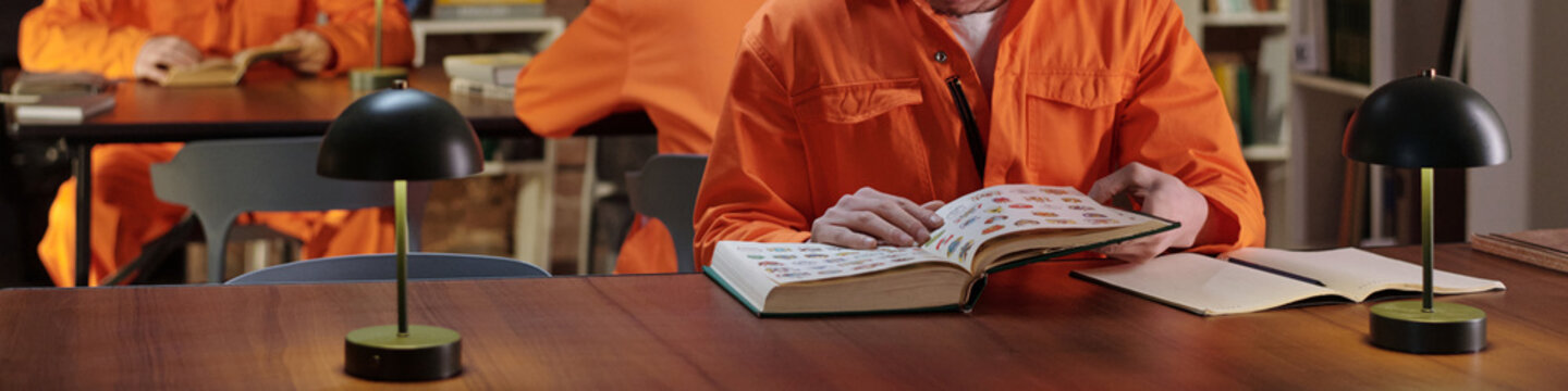 Caucasian man reading large book at table in prison library, other inmates in orange uniforms studying in background, bookshelves visible, educational setting for incarcerated individuals