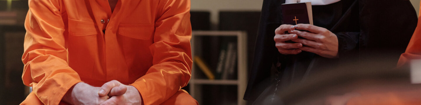 Caucasian man in orange prison uniform sitting next to Caucasian woman dressed as nun holding book with cross, both seated in prison library setting
