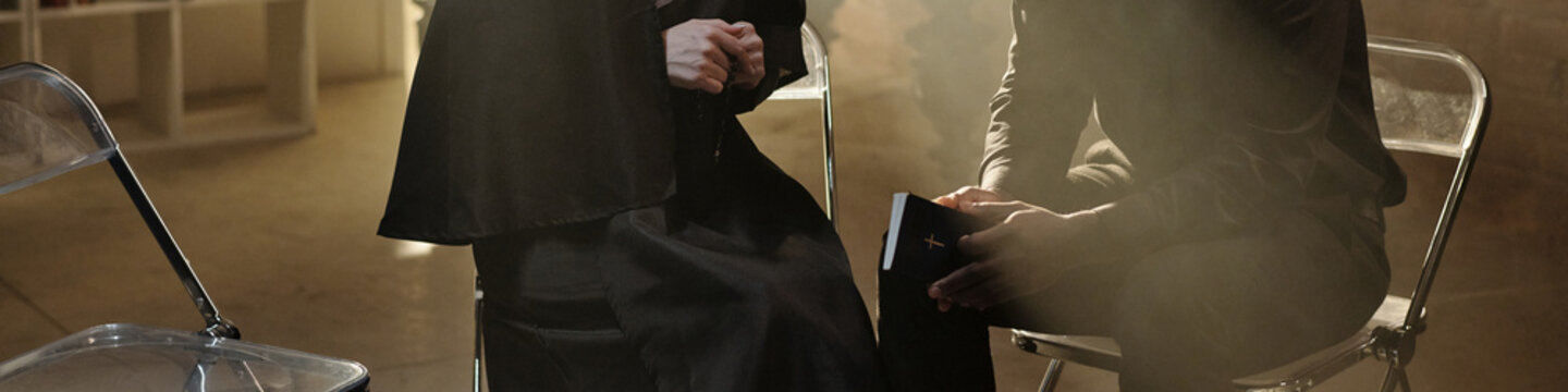 Caucasian woman dressed as nun sitting beside Black priest in prison library engaging in conversation with bookshelves visible in background