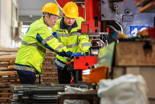 Two male industrial workers wea safety helmets and reflective jackets operating large industrial machinery in a factory setting with tools and equipment around