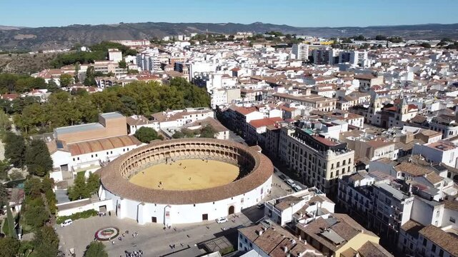 Drone shot of circular bullring in center of town with surrounding white buildings, narrow streets, and distant hills, capturing contrast between architecture and natural terrain