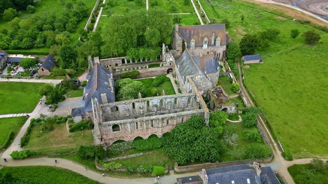 Drone shot pulling back from Beauport Abbey ruins, revealing gardens, paths, and the low tide beach - Frabce