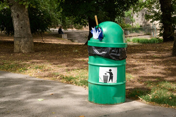 A green trash can in a park, partly covered with a black bag and holding an umbrella, is surrounded...
