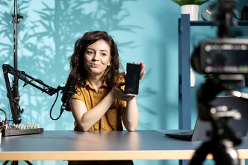 Cheerful entertainer presenting mobile phone from brand sponsoring video, urging viewers to...
