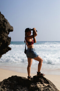 Woman traveler with camera taking pictures of ocean from rock on beach