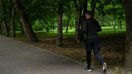 Man jogs along a pathway in a lush green park surrounded by trees during midday