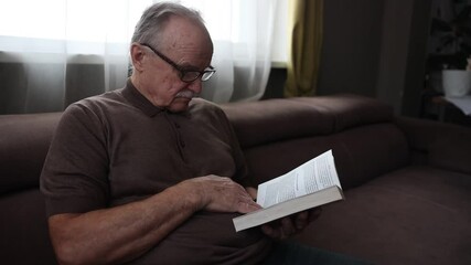 senior man with glasses reads a book while sitting on the couch at home.  Slow life. Enjoying the little things. fighting dementia