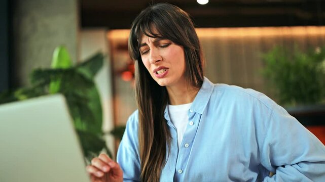 Sick unwell girl experiences severe stomach pain and discomfort working on laptop in a contemporary office. Dressed in light blue shirt, she struggles with chronic illness, seeking relief and coping.