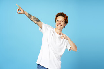 Cheerful individual with short red hair and tattoos strikes a playful pose, pointing upward