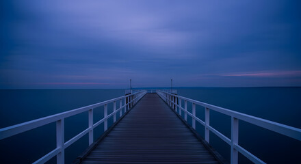 Fototapeta premium Long empty pier with white railings extends into the calm deep blue sea at twilight