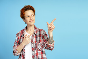 Portrait of optimistic woman with red hair, piercing and tattoo on arm, pointing to copy space