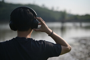 A person wearing large headphones listens to audio while standing outdoors near a calm body of...