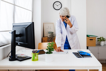 Senior female doctor talking on phone with patient while using desktop computer to check his history standing at desk in hospital