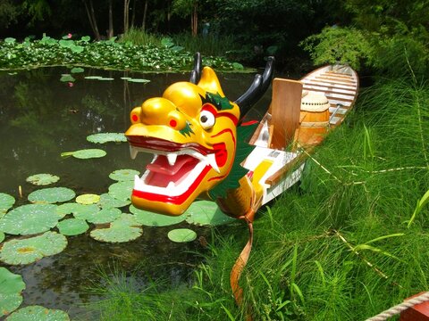 A colorful, carved dragon boat rests by the shore of a forested, lotus-flower lake.