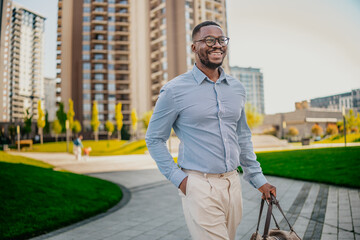 Black businessman walking confidently in urban park setting with buildings in background during...