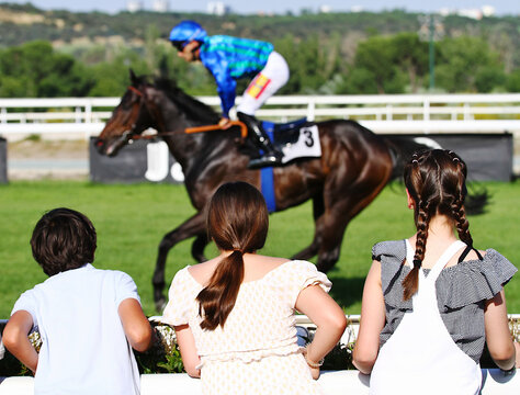 P&uacute;blico espectador de hip&oacute;dromo con ni&ntilde;os de espaldas mirando atentos caballo galopando con jockey en pista de hierba turf