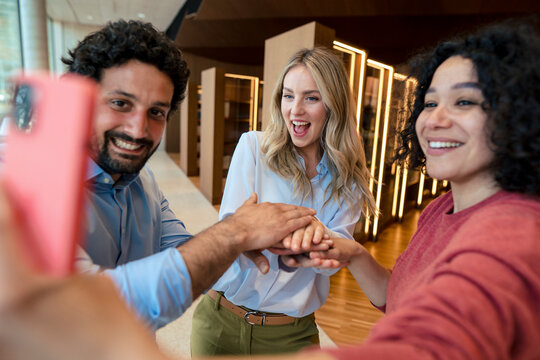 Happy colleagues stacking hands and taking selfie on smart phone in library