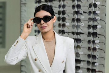 Attractive Caucasian young brunette woman in white suit trying on and choosing sunglasses to buy at...