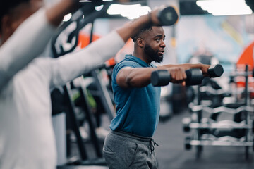 Determined black man lifting dumbbells in gym with people exercising