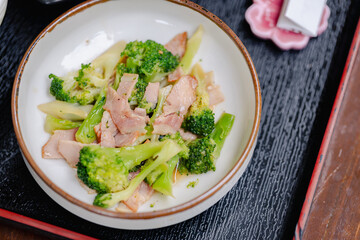 Stir fried broccoli with slices of cooked meat served in ceramic bowl on textured black tray, offering healthy and delicious meal option with fresh vegetables and protein