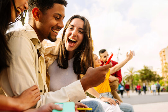 Young adult friends using mobile phone together outside. Gen Z people generation having fun watching social media content on cell. Youth and technology concept