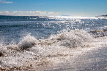 Strong waves break on the shore while the sun reflects on the golden surface of the sea.