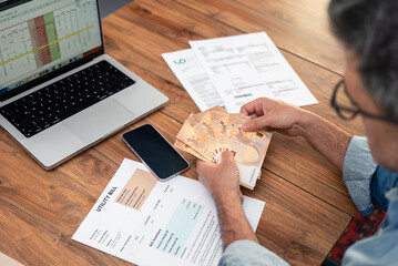 Senior male tenant holds euros fan at wooden table with utility bills, smartphone and laptop,...