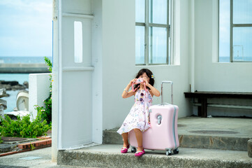 Young Japanese girl taking a picture with a toy camera next to a pink suitcase at a seaside bus...