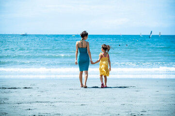 A smiling Japanese woman and her daughter holding hands and walking together at the edge of the sea...