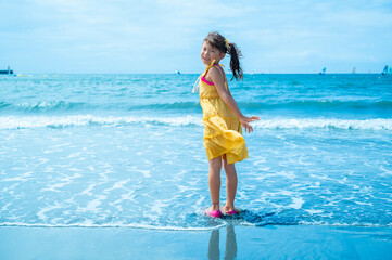 Young Japanese girl in a yellow dress playing joyfully in the ocean waves on a bright summer day,...