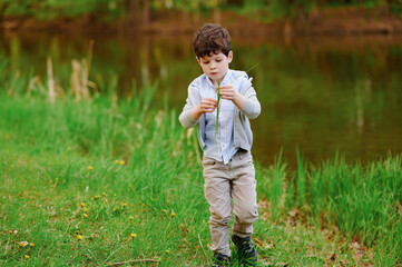 young boy exploring nature by a lakeside holding grass stems, wearing casual clothing. capturing...