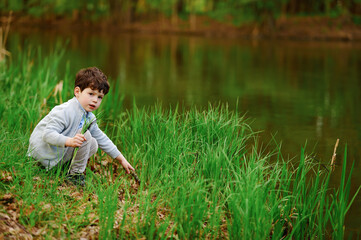 curious child exploring lakeside nature, crouching by water's edge. young boy in casual clothing,...