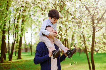 father and son enjoying playful moment outdoors in blooming park. parent carrying child on shoulders, surrounded by spring blossoms and lush greenery, symbolizing family joy and bonding