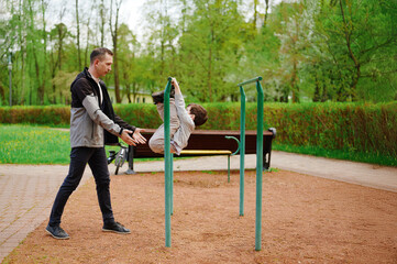 father supervising son at playground. father supports young boy performing gymnastics on outdoor...