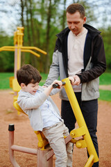 Obraz premium father and son enjoying outdoor fitness equipment in park. man guiding child on yellow exercise machine. family time promotes healthy lifestyle and bonding moments in nature