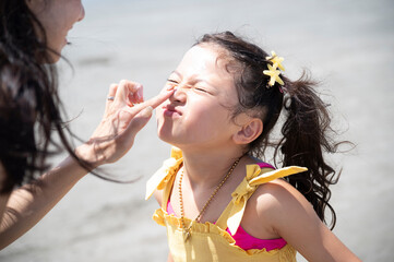 Mother gently applying sunscreen to her daughter's cheeks during a fun summer day at the beach,...