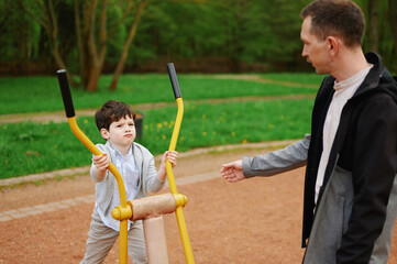 father and son exercising on outdoor fitness equipment in park. child gripping handles with...