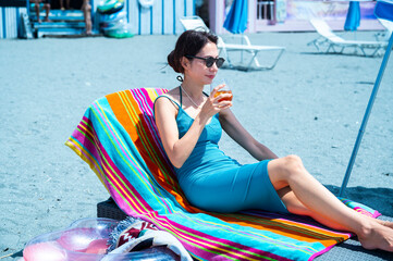 Japanese mother reclining on a colourful striped towel-covered beach lounger, smiling and enjoying...