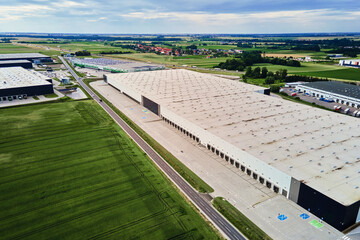 Large modern warehouse with loading docks and parked trucks surrounded by green fields. Industrial...