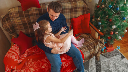 Happy family having fun at home for Christmas. Father tickles her daughter lying on the sofa....