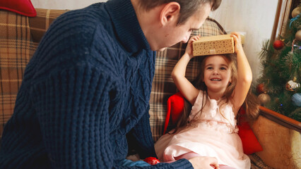 Dad is giving a gift box to his beautiful little daughter. Near the Christmas tree at home