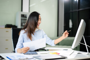 Happy Asian businesswoman reviews financial reports at her office desk. Bright workspace,...