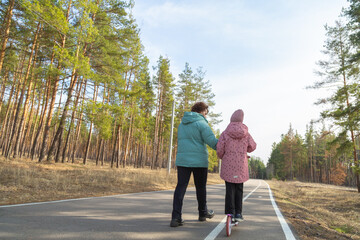 Little girl riding scooter while walking with her grandmother in the forest on a sunny day